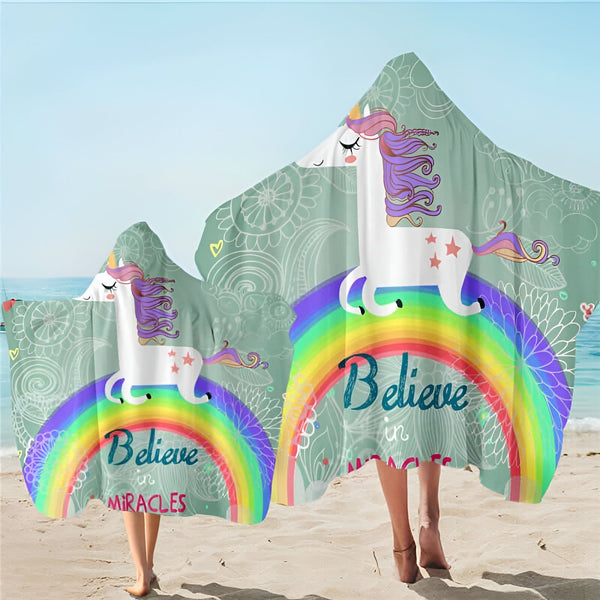 Deux personnes sur une plage, portant des serviettes à capuche avec un motif de licorne sur un arc-en-ciel, le texte « Believe in Miracles » écrit en dessous, sur un fond vert d'eau., 45501252108543 | Licorne Shop