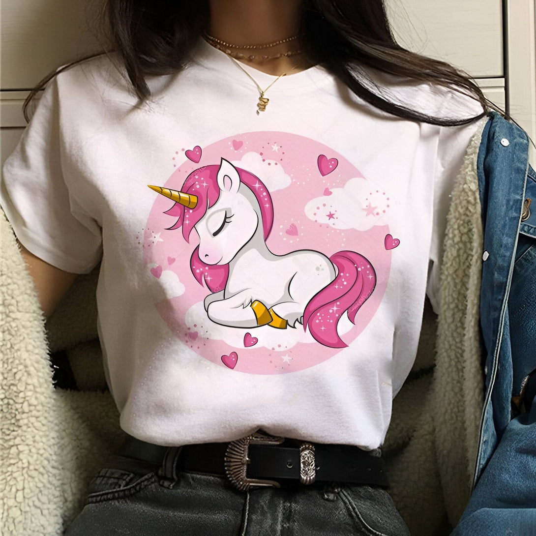 Une jeune femme porte un t-shirt blanc avec une image d'une licorne endormie sur un nuage. La licorne a une crinière et une queue roses et une corne dorée. Il y a des coeurs roses et des nuages blancs autour de la licorne.