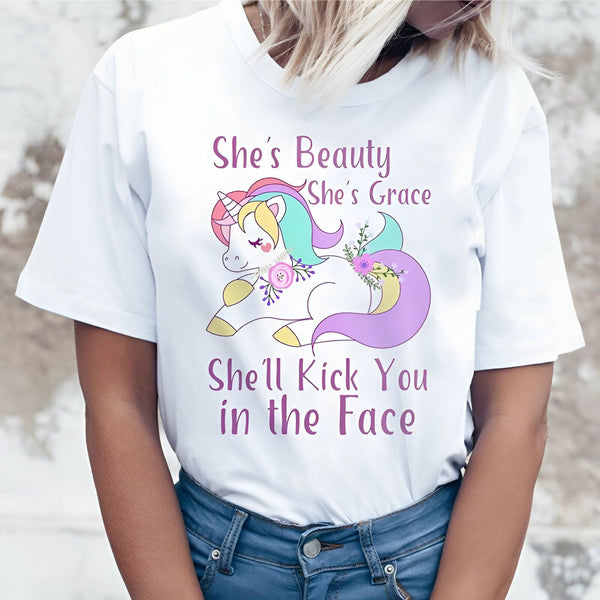 Une image d'une licorne blanche avec une crinière et une queue arc-en-ciel. La licorne est allongée sur le sol et porte des fleurs sur la tête et la queue. Le texte sur l'image dit "She's beauty, she's grace, she'll kick you in the face".