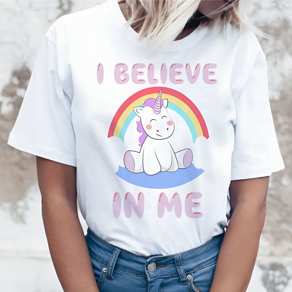 Une jeune femme porte un t-shirt blanc avec un arc-en-ciel et une licorne. La licorne est assise sur un nuage et le texte "I believe in me" est écrit en dessous.