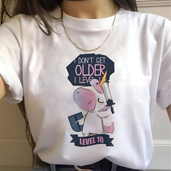 Une femme porte un t-shirt blanc avec un dessin d'une licorne tenant une épée et un bouclier. Il est écrit sur le t-shirt "I don't get older, I level up" et "Level 10".