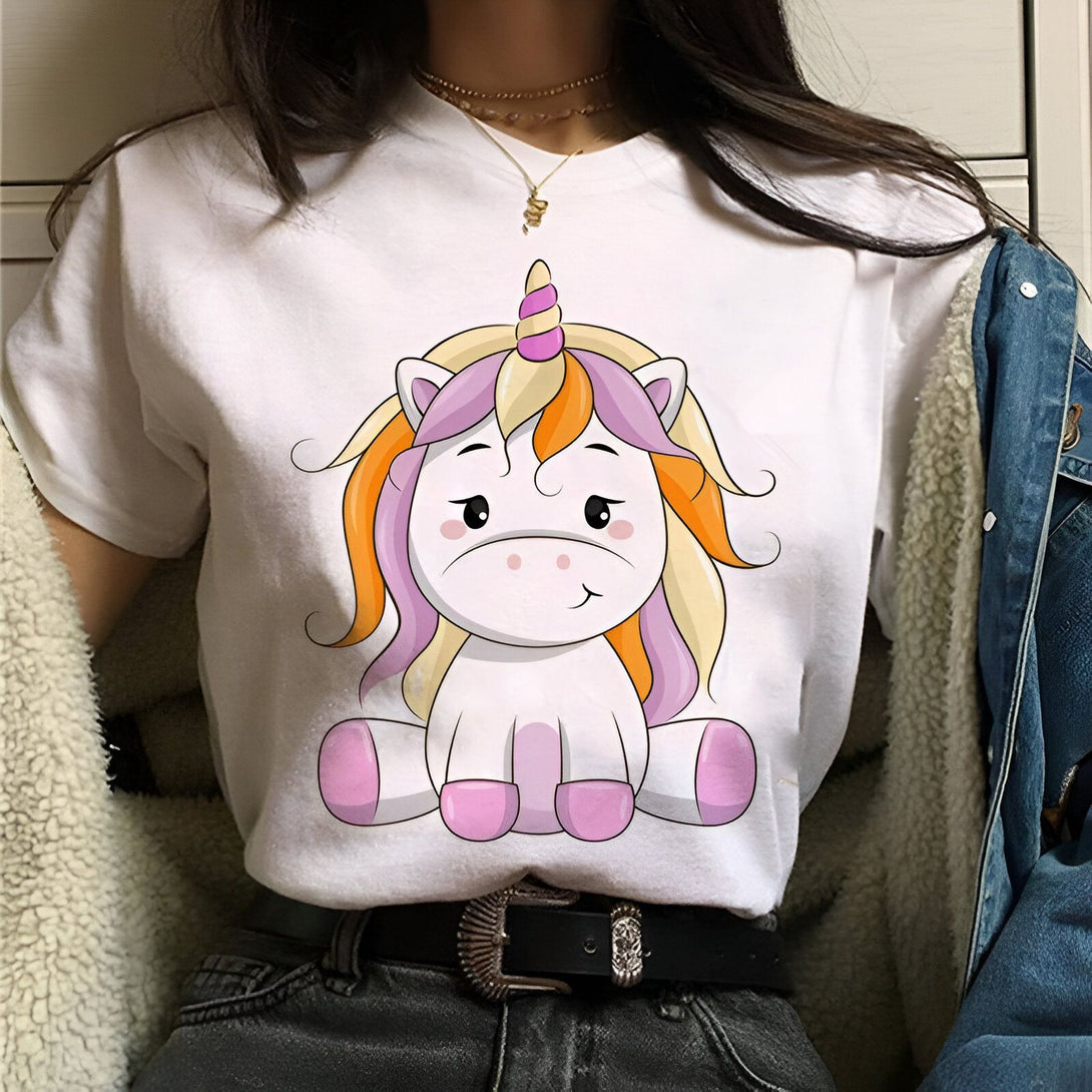 Une jeune femme porte un t-shirt blanc avec une image de licorne. La licorne est assise et a une crinière et une queue multicolores. La femme a un jean bleu et une veste en jean bleue.