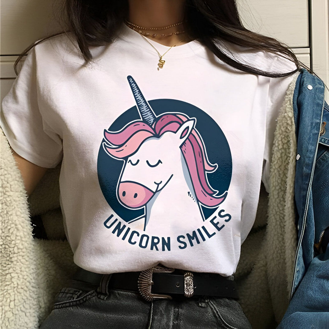 Une jeune femme porte un t-shirt blanc avec un dessin de licorne. La licorne a une crinière et une queue roses. Le texte "Unicorn Smiles" est écrit sous la licorne.