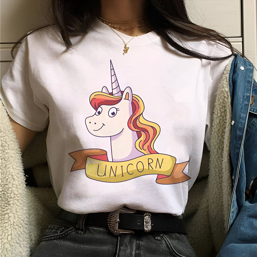 Une jeune femme porte un t-shirt blanc avec un dessin de licorne. La licorne a une crinière et une queue arc-en-ciel. Le texte "UNICORN" est écrit en lettres jaunes sur une banderole rose.