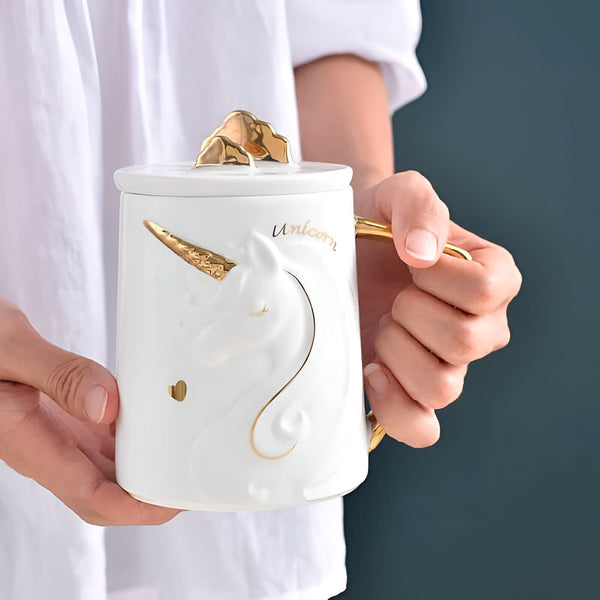 Gros plan d'une personne tenant une tasse en céramique blanche ornée d'un motif de licorne en relief doré, avec un couvercle surmonté d'une décoration dorée en forme de nuage., 45501192798463 | Licorne Shop