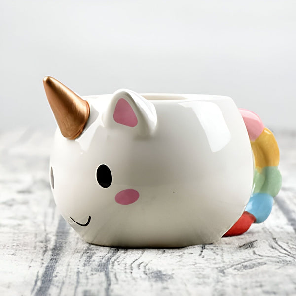 Tasse en céramique en forme de licorne blanche avec corne dorée et queue arc-en-ciel, yeux noirs et joues roses, sur une surface en bois., 45501197648127 | Licorne Shop