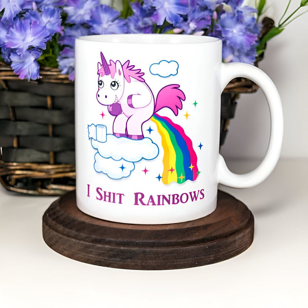 Mug blanc avec un dessin humoristique de licorne rose faisant des arcs-en-ciel sur un nuage. Inscription "I Shit Rainbows". Fleurs bleues en arrière-plan. Idée cadeau drôle., 45501199220991 | Licorne Shop