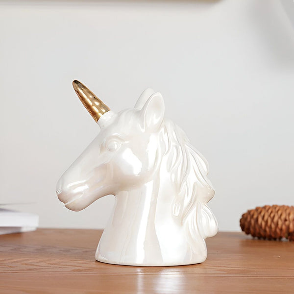 Une sculpture en céramique d'une tête de licorne blanche avec une corne dorée. La sculpture est posée sur une surface en bois.