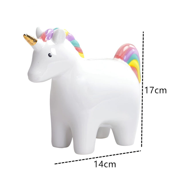 Une tirelire en céramique blanche en forme de licorne. La licorne a une crinière et une queue arc-en-ciel et une corne dorée. La tirelire mesure 17 cm de haut et 14 cm de large.