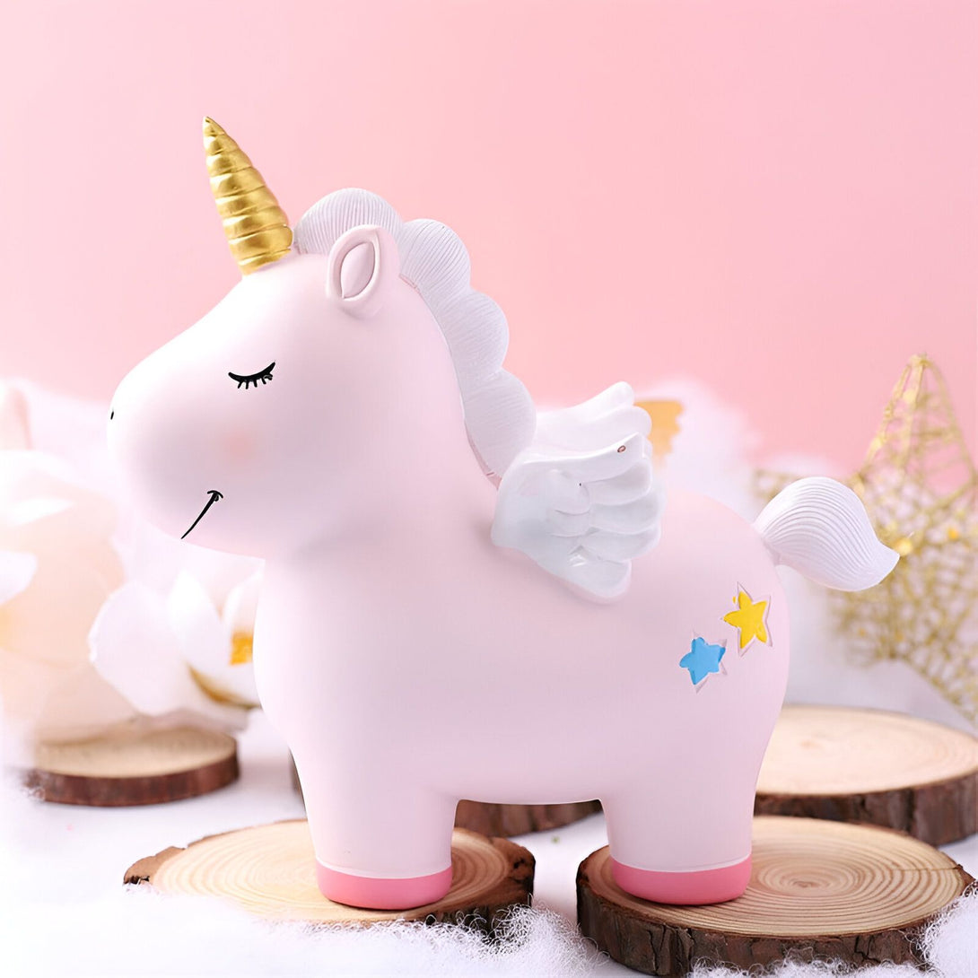 Une tirelire en forme de licorne rose avec une crinière et une queue blanches. La licorne a une corne dorée et des ailes bleues. Elle est posée sur une bûche de bois.