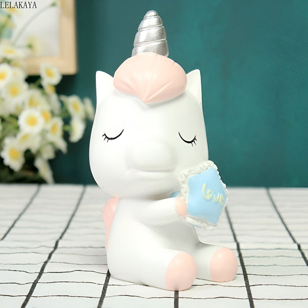 Une figurine de licorne blanche et rose tenant un oreiller bleu avec le mot "love" dessus. La licorne a les yeux fermés et un sourire sur le visage. Elle est assise sur une table à carreaux noirs et blancs. En arrière-plan, on peut apercevoir un mur vert et des fleurs blanches.