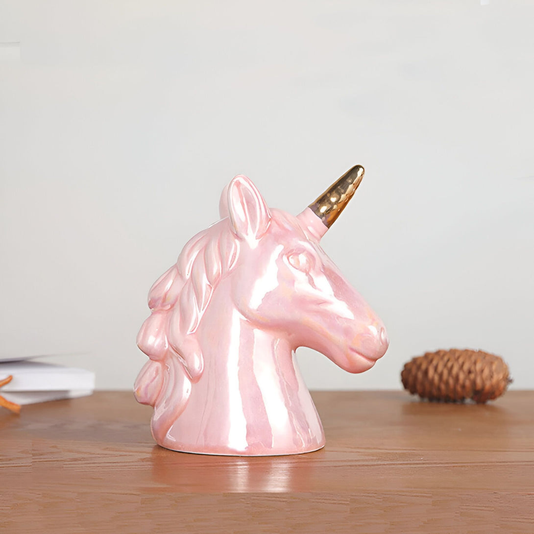 Une sculpture en céramique d'une tête de licorne rose avec une corne dorée. La sculpture est posée sur une surface en bois.