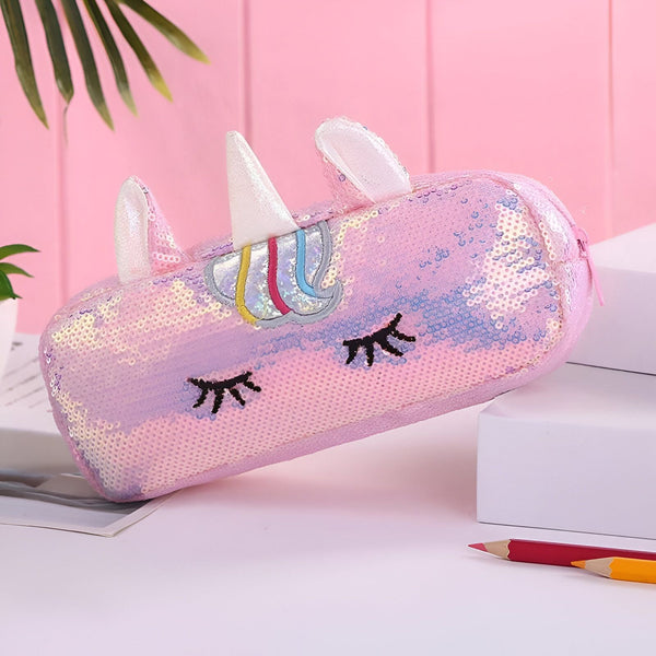 L'image montre une trousse rose en forme de licorne. La trousse est recouverte de paillettes et a une fermeture éclair. La licorne a une crinière et une queue arc-en-ciel, ainsi que des yeux et des oreilles en 3D.