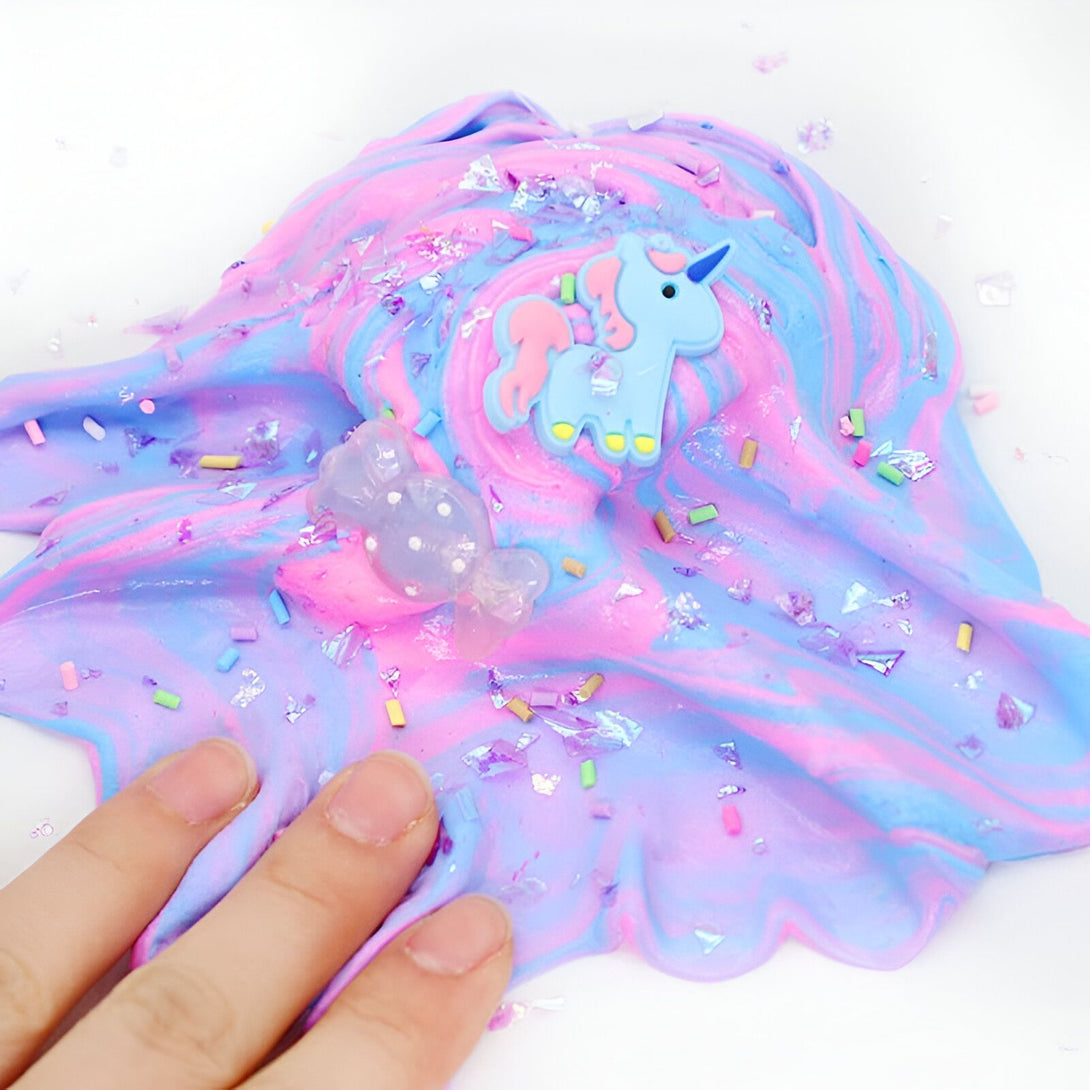 Gros plan d'une main manipulant une pâte à modeler (slime) aux couleurs pastel bleu, rose et violet. Décorations : licorne, bonbon transparent et paillettes multicolores., 45501394845951 | Licorne Shop
