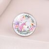 bague-licorne-design-3a L'image représente un bouton de meuble rond en métal argenté avec un cabochon en verre bombé. Sur le cabochon, on voit une illustration d'une licorne blanche avec une crinière et une queue roses. La licorne est entourée d'un cercle de fleurs roses, mauves et jaunes.