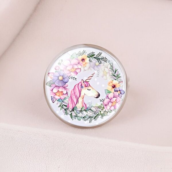 L'image représente un bouton de meuble rond en métal argenté avec un cabochon en verre bombé. Sur le cabochon, on voit une illustration d'une licorne blanche avec une crinière et une queue roses. La licorne est entourée d'un cercle de fleurs roses, mauves et jaunes.