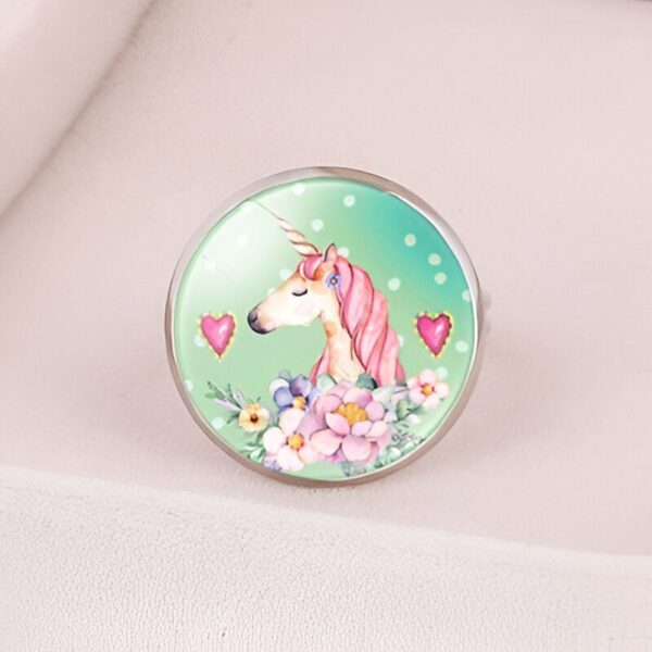 L'image représente une bague en acier inoxydable avec un cabochon en verre rond. Le cabochon est décoré d'une illustration d'une licorne blanche avec une crinière et une queue roses. La licorne est entourée de fleurs roses et vertes.