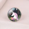 L'image représente une bague en métal argenté avec un cabochon en verre rond. Le cabochon est décoré d'une illustration colorée représentant une licorne blanche avec une crinière et une queue roses et violettes. La licorne est entourée de fleurs et de feuillage.