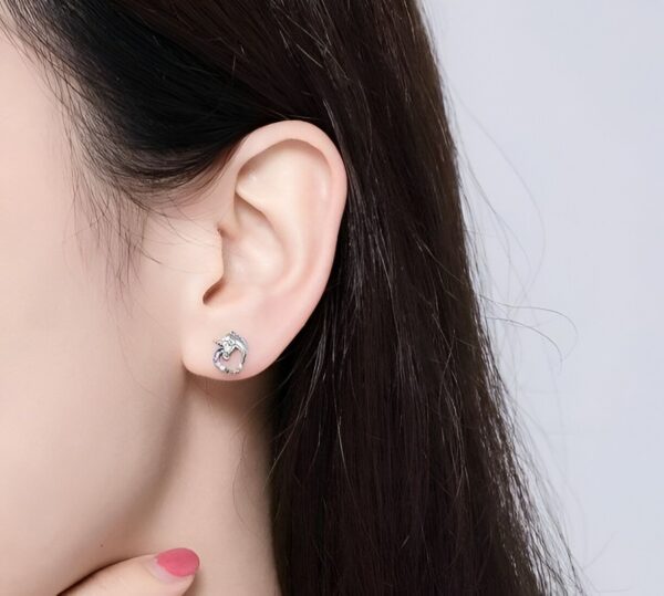 boucles-d-oreilles-licorne-diamant-argent-2a.jpg Une image d'une femme portant des boucles d'oreilles en forme de licorne en argent sterling. Les boucles d'oreilles sont serties de zircons cubiques transparents et ont une finition polie. La femme a les cheveux longs et noirs et porte un maquillage naturel. Elle sourit et regarde l'appareil photo.