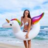 Une jeune femme souriante en bikini et lunettes de soleil, sur la plage, posant avec une bouée gonflable en forme de licorne blanche avec une crinière arc-en-ciel et des ailes colorées. Ambiance estivale et vacances., 45501249421567 | Licorne Shop