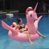 bouee-geante-licorne-2-places-1a.jpg Une femme en bikini se détend sur une bouée gonflable rose en forme de licorne dans une piscine. Le ciel est dégagé et l'eau est bleue., 45501251682559 | Licorne Shop