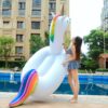 Une jeune femme en maillot de bain rayé se tient à côté d'une grande bouée licorne gonflable près d'une piscine. La bouée est blanche avec une crinière et une queue arc-en-ciel, et une corne dorée., 45501249847551 | Licorne Shop