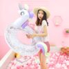 bouee-gonflable-licorne-geante-1a.jpg Une jeune femme en chapeau de paille tient une bouée gonflable licorne scintillante dans un décor rose pastel avec des balles roses et blanches., 45501244244223 | Licorne Shop