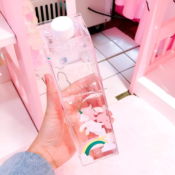 bouteille-d-eau-licorne-en-plastique-pour-enfant-1a.jpg Une main tient une bouteille d'eau transparente en forme de brique de lait avec un bouchon blanc. Des dessins d'arcs-en-ciel et de licornes décorent la bouteille. L'arrière-plan est rose., 45501173858559 | Licorne Shop