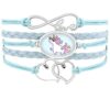L'image montre un bracelet en cuir bleu avec un pendentif en forme de licorne. La licorne est représentée avec des lunettes de soleil et une crinière arc-en-ciel. Le bracelet est également décoré de deux cœurs en métal.