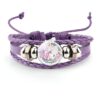 bracelet-licorne-enfant-tresse-1a-1.jpg Le bracelet en cuir tressé de couleur mauve comporte un médaillon rond en verre avec une image de licorne blanche avec une crinière et une queue roses et des fleurs colorées tout autour.