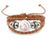 bracelet-licorne-enfant-tresse-4a.jpg Un bracelet en cuir marron avec un pendentif en verre rond. Le pendentif représente une licorne blanche avec une crinière et une queue roses. La licorne est entourée de fleurs roses et vertes.