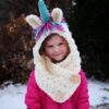 capuche-echarpe-licorne-chaude-pour-enfant-1a.jpg Fillette souriante portant un bonnet/écharpe en crochet blanc en forme de licorne avec une corne bleue, des oreilles blanches et des mèches de fil rose et bleu. Elle porte une veste rose vif., 45502093721855 | Licorne Shop