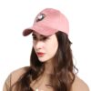 Une jeune femme porte une casquette rose en daim avec une broderie d'une licorne. La licorne a une crinière arc-en-ciel et un œil fermé. La femme a les cheveux longs et bruns et porte un maquillage léger. Elle regarde vers le bas.
