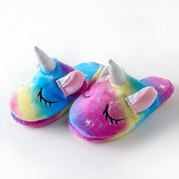 Une paire de pantoufles licorne en peluche arc-en-ciel avec une corne et des oreilles argentées. Les pantoufles sont fermées et ont une semelle antidérapante.