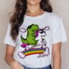 Femme portant un T-shirt blanc avec un dinosaure vert et une licorne blanche, tous deux portant des lunettes de soleil, chevauchant un arc-en-ciel avec des nuages. Colliers dorés à son cou., 45502330274047,45502330306815,45502330339583,45502330372351,45502330405119,45502330437887,45502330470655,45502330503423 | Licorne Shop