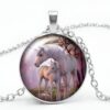 collier-licorne-mere-et-fille-1a.jpg Un pendentif rond en métal argenté avec une chaîne. Le pendentif représente une licorne blanche et son petit dans une forêt. La licorne et son petit sont debout sur un sol vert avec des arbres et des montagnes en arrière-plan. Le ciel est rose et il y a un arc-en-ciel qui traverse le ciel.