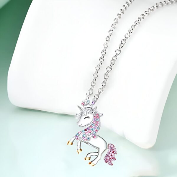 collier-licorne-princesse-2a.jpg L'image représente un collier avec un pendentif en forme de licorne. La licorne est blanche avec une crinière et une queue roses et bleues. Elle a une corne dorée et un collier avec des pierres précieuses roses et bleues. Le collier est en argent.