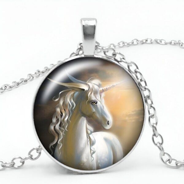 Un pendentif rond en métal argenté avec une chaîne. Le pendentif représente une licorne blanche avec une crinière et une queue arc-en-ciel. La licorne est debout dans un pré vert avec des fleurs. Le ciel est bleu avec des nuages blancs.