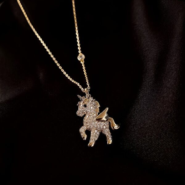 collier-licorne-strass-or-1a.jpg Un pendentif en forme de licorne en or avec des ailes et des pierres précieuses sur un fond noir.
