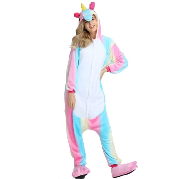 combinaison-licorne-multicolore-adulte-1a.jpg Une femme porte un pyjama licorne. Le pyjama est rose, bleu et blanc. Il a une capuche avec une corne de licorne. La femme est heureuse et sourit.