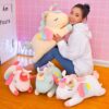 Une femme souriante serre un grand doudou licorne jaune. Autour d'elle, plusieurs autres peluches licornes colorées sont posées sur le sol. Décor rose et inscription lumineuse en arrière-plan., 45501048684799 | Licorne Shop