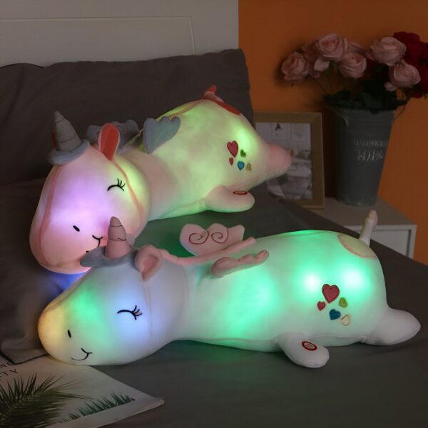 coussin-lumineux-licorne-veilleuse-1a.jpg Deux peluches licornes lumineuses reposent sur un lit. Elles sont colorées et ont des motifs de cœurs. Des fleurs décorent l'arrière-plan., 45501066281215 | Licorne Shop