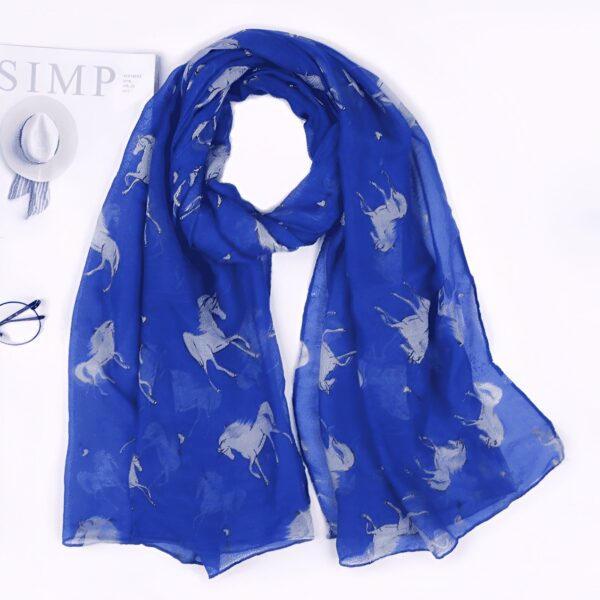 echarpe-bleue-licorne-a-corne-et-pompons-pour-femme-1a.jpg Écharpe bleu roi à motifs de chevaux argentés, posée à plat près d'un chapeau, de lunettes et d'un magazine. Accessoire de mode élégant et tendance pour femme., 45502083825919 | Licorne Shop