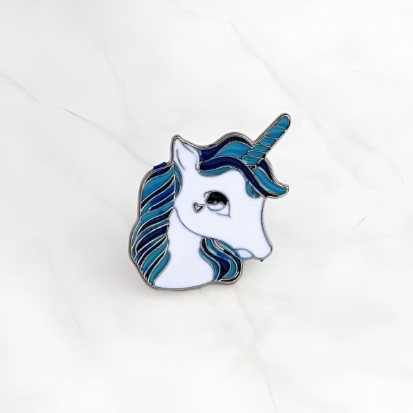 Pin's en émail représentant la tête d'une licorne blanche avec une crinière et une corne bleues, avec un petit cœur dessiné à la place de l'œil., 45502390894847 | Licorne Shop