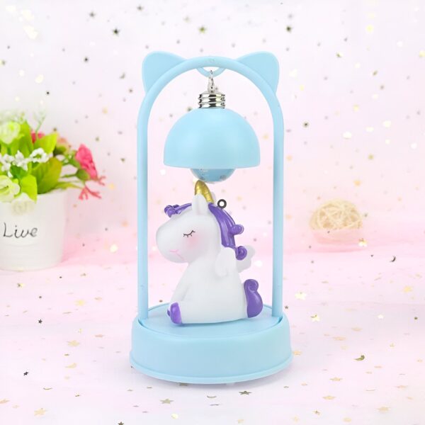 lampe-de-chevet-licorne-bleue-1a.jpg Une veilleuse en forme de licorne blanche et violette avec une crinière et une queue arc-en-ciel. La licorne est assise sur un nuage bleu et est entourée d'étoiles. La veilleuse est posée sur un socle bleu.
