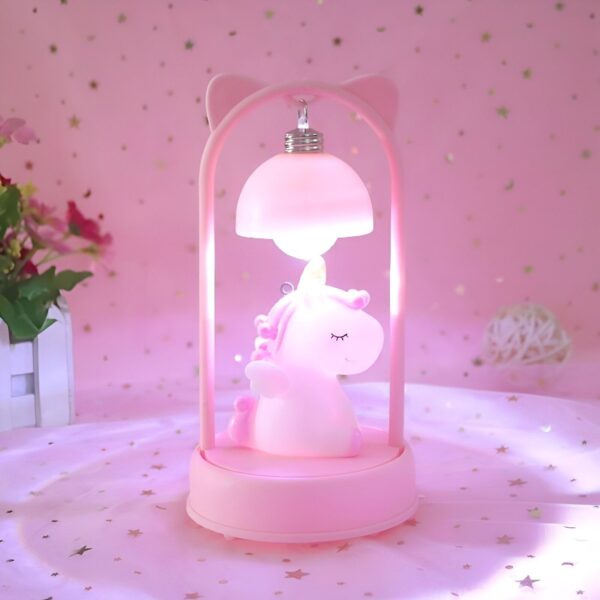 lampe-de-chevet-licorne-rose-1a.jpg Une veilleuse rose en forme de licorne avec un abat-jour en forme de champignon. La licorne est allongée et a les yeux fermés. La veilleuse est posée sur une table rose.