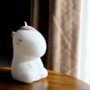 Une veilleuse licorne blanche en forme de dessin animé est posée sur une table en bois. La licorne a une crinière et une queue roses et bleues. Elle est endormie et a les yeux fermés.