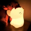 Une veilleuse en forme de licorne blanche avec une corne et une crinière roses. La veilleuse est allumée et émet une lumière douce.