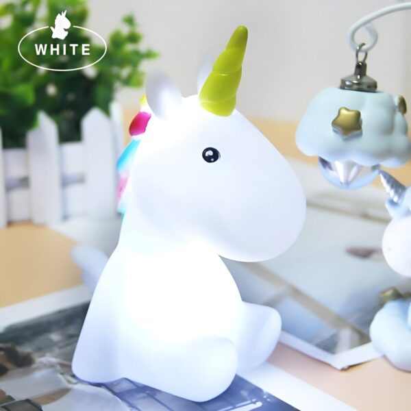 lampe-veilleuse-licorne-1a.jpg Une veilleuse en forme de licorne blanche avec une corne jaune et une crinière multicolore. Elle est assise sur une table avec un fond blanc.