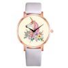 Une montre blanche avec un cadran rond et un bracelet en cuir blanc. Le cadran représente une licorne blanche avec une crinière et une queue roses. La licorne est entourée de fleurs roses, jaunes et bleues.